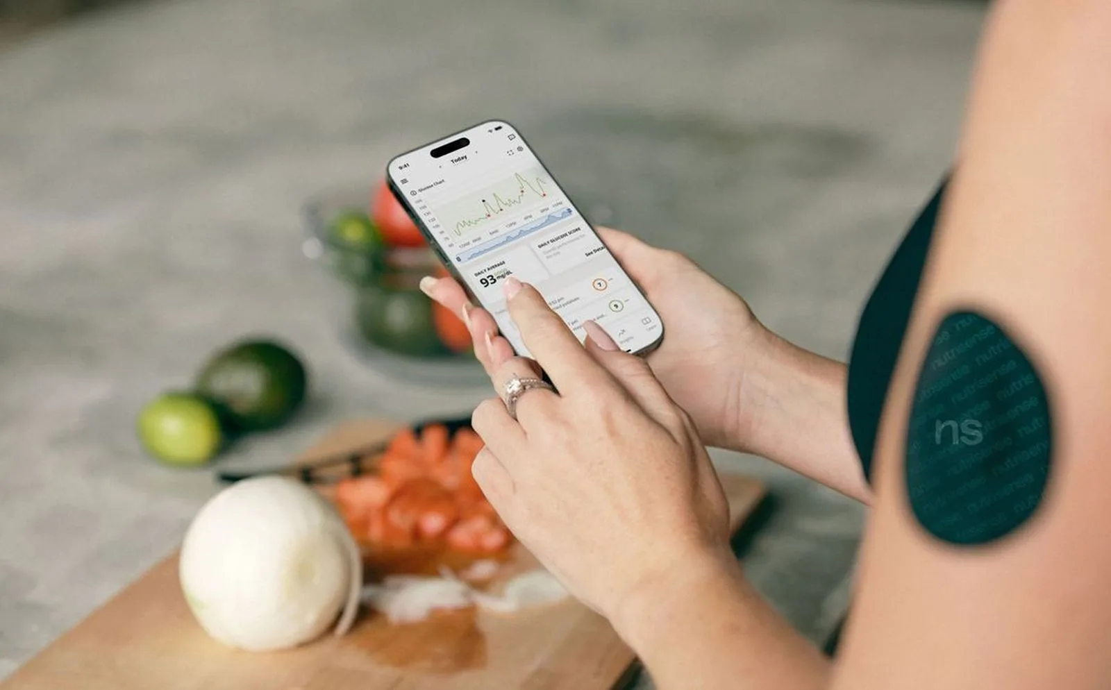 A woman wearing a continuous glucose monitor checks glucose information on her phone while preparing a meal.